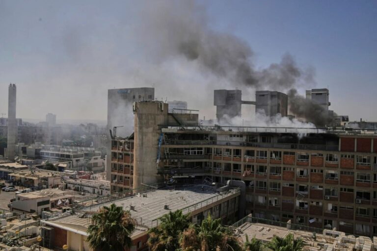 The aftermath of a missile strike on Soroka Hospital in Beersheba. Dark smoke pours from the damaged upper floors of the medical center, with a hazy, smoke-filled sky in the background.