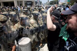 A man in a black shirt and baseball cap gestures with his hand up in front of a line of California National Guard soldiers in camouflage uniforms and riot gear, including helmets with face shields and large riot shields. A crowd of people is visible behind them on a city street during the day.