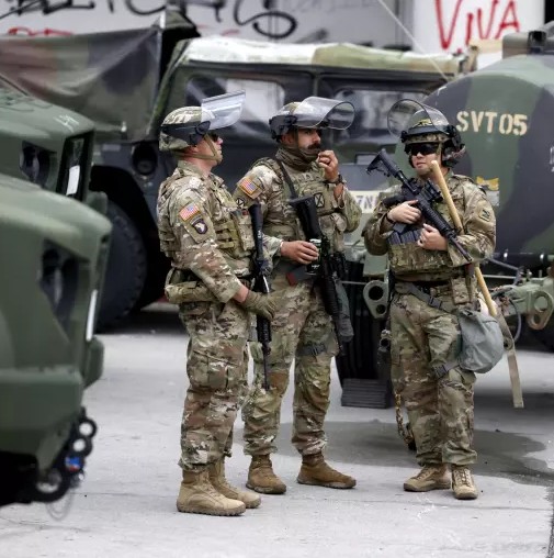 California National Guard troops in camouflage uniforms and riot gear stand guard on a city street in Los Angeles.